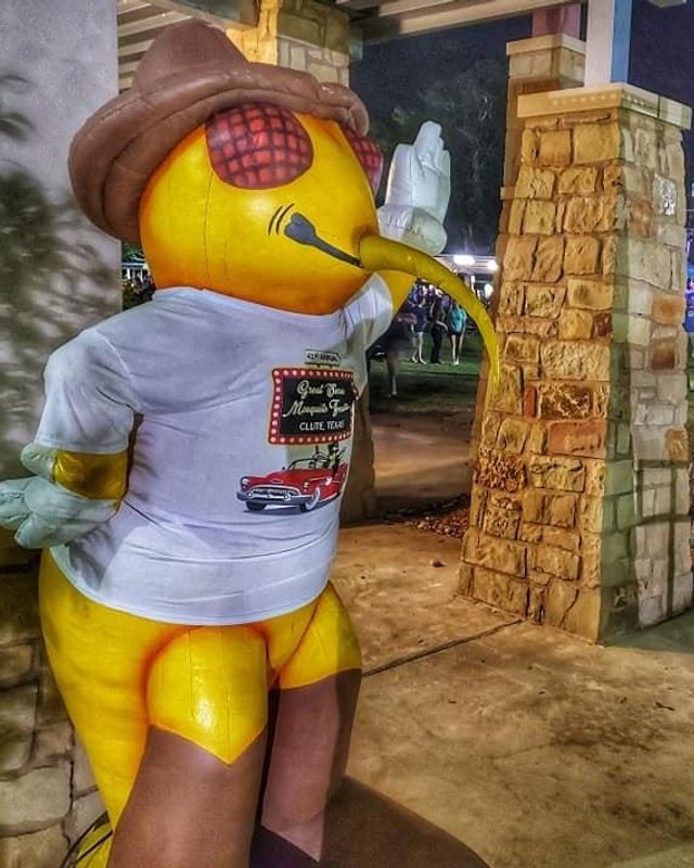 Giant yellow mosquito mascot in a hat and t-shirt at the festival grounds at night, with a crowd in the background.