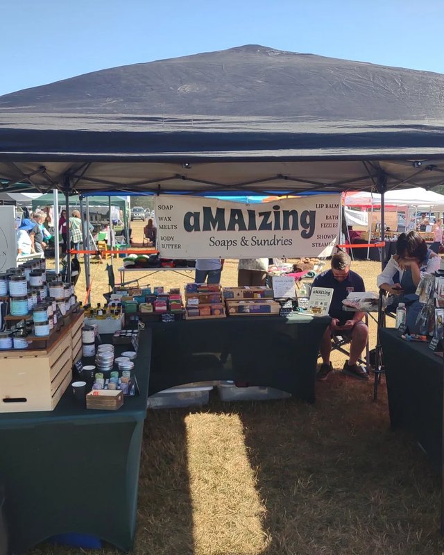 Vendors at the aMAIZing Soaps and Sundries booth, with soap jars and goods displayed under a canopy at an outdoor fair.