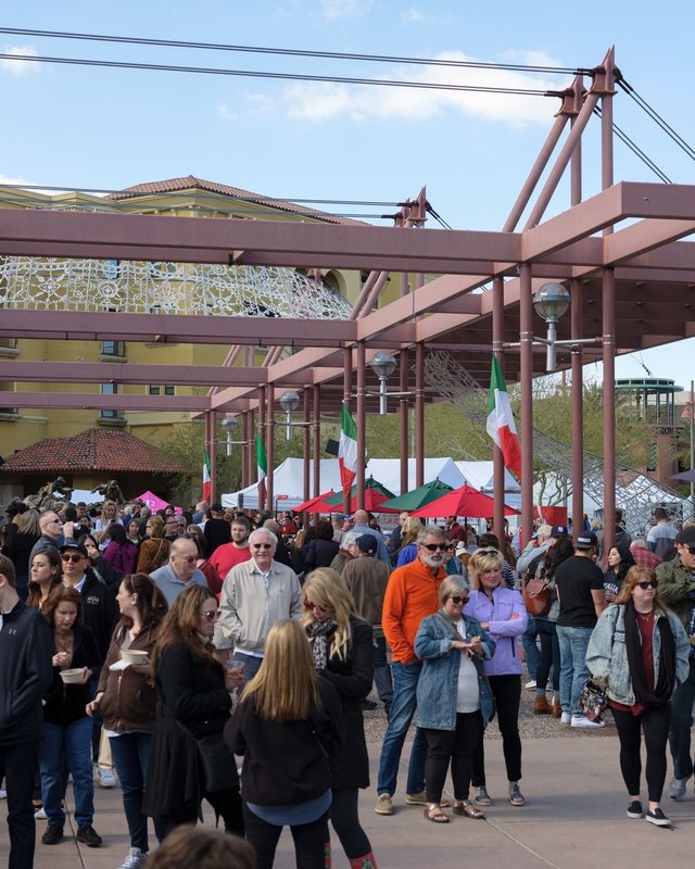 Festival crowd walking past white tents under a steel pergola at the Scottsdale Italian Festival.