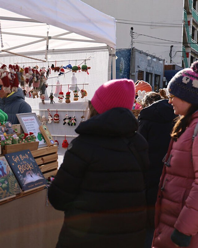 Vendors at a holiday market stall selling ornaments and gifts under a white canopy, with shoppers browsing.