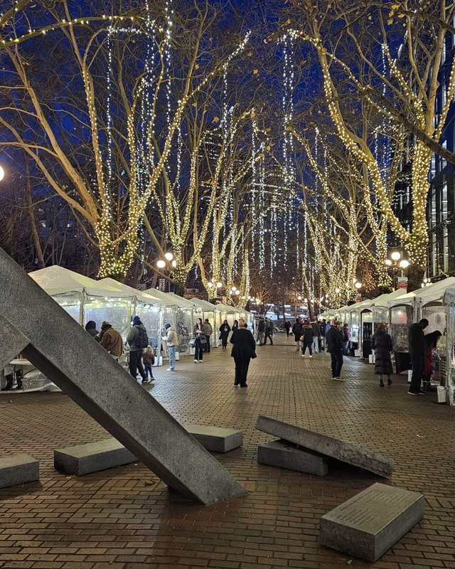 Holiday lights over tents and shoppers along a Pioneer Square street at night