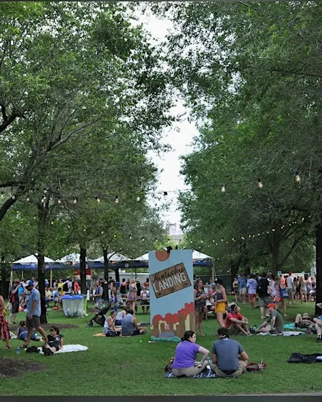 Outdoor park fair with white vendor tents under tall trees and a crowd of visitors on the lawn.