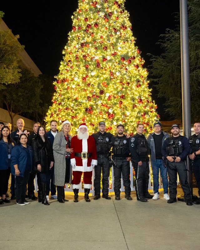 Group of city staff and officers posing with Santa in front of a large lit holiday tree at night.