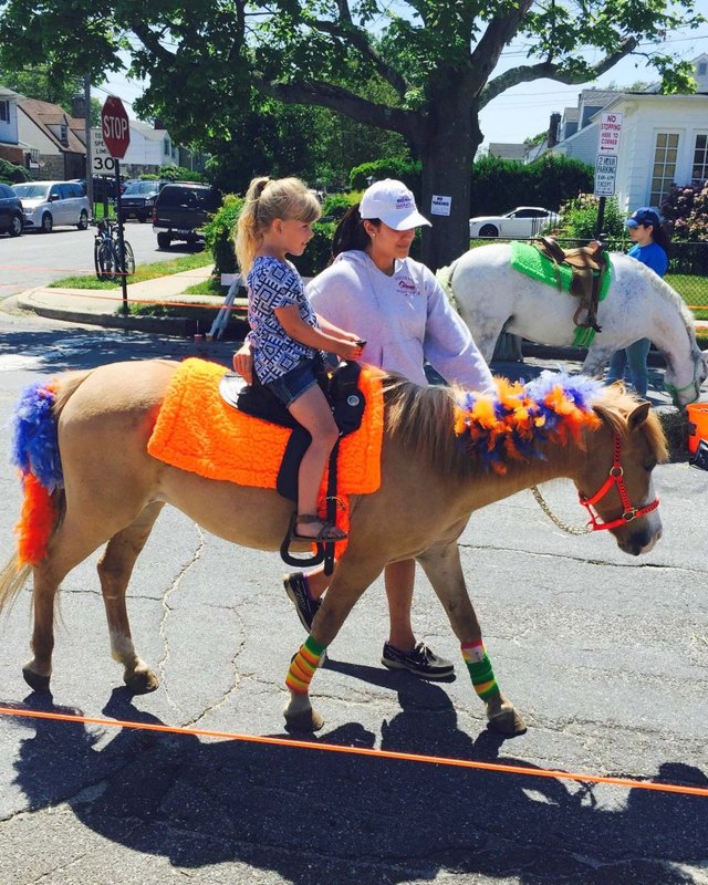 A child and an adult riding a pony decorated with orange and blue trim on a street lined with trees and houses.