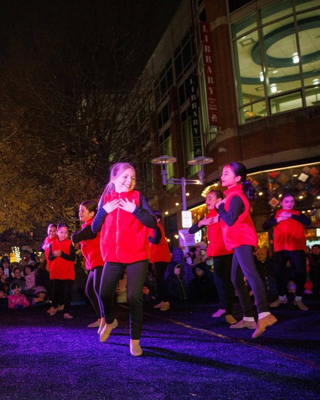 Performers in matching red vests dance on purple-lit ground in front of a crowd at night, with a multi-story building behind them.