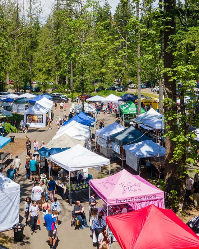 Aerial view of the Maple Valley Farmers Market with white and pink booth tents and shoppers under trees.