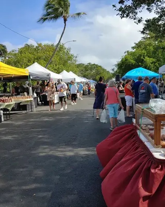 Market aisle with canopy booths, produce tables and shoppers browsing at the Oceanside farmers market in Lake Worth Beach.