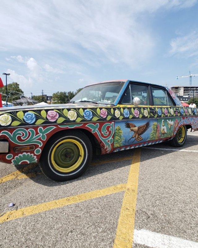 Decorated vintage car with painted flowers and birds parked on pavement at Louisville Maker Faire.