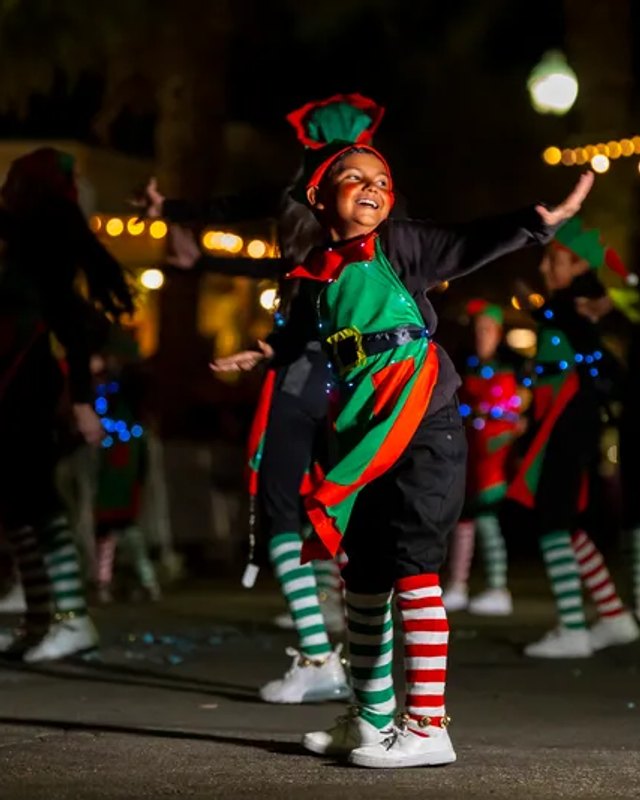 Performer in an elf costume dancing in front of a line of holiday lights at a night parade in Coachella.