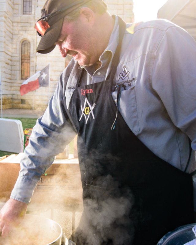Man in a cap and apron with Masonic emblems leans over a steaming pot at a festival booth; a Texas flag and stone building are behind him.