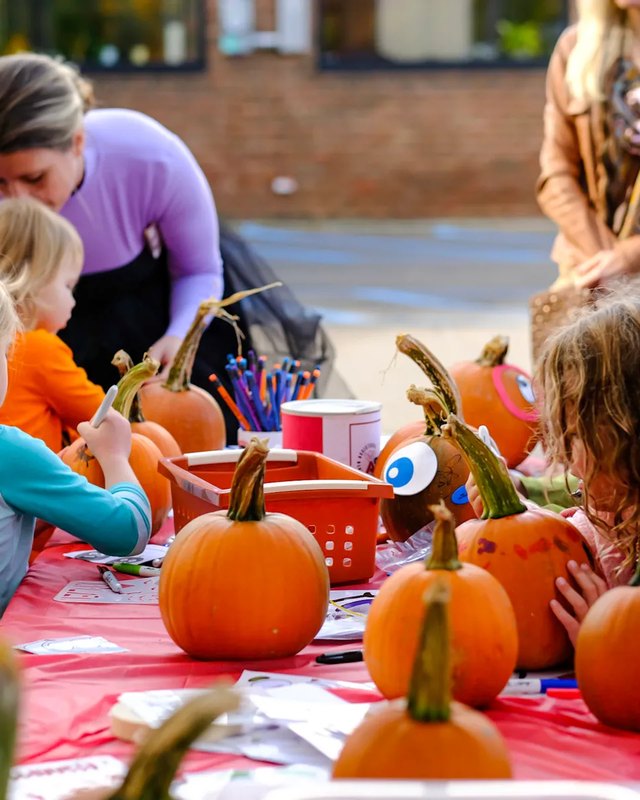 Children decorating pumpkins at a red-covered table at an outdoor fall market event.