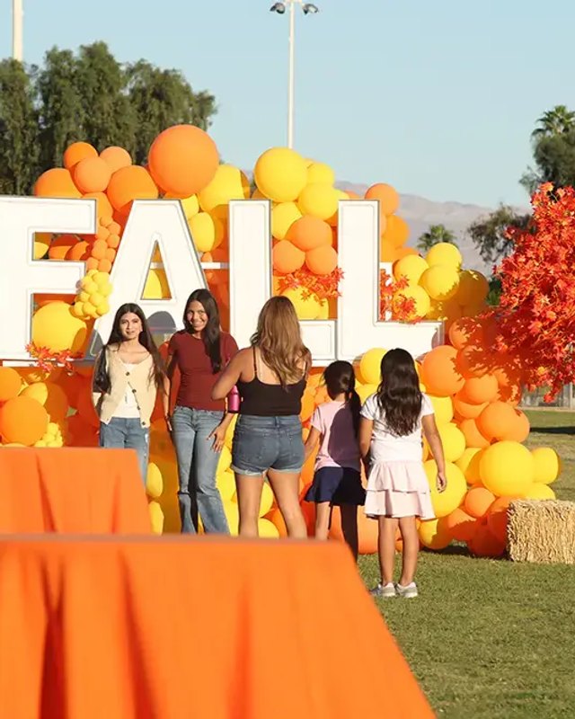 Young women and children posing in front of a large yellow and orange balloon arch spelling FALL, with orange tables, at a park event.