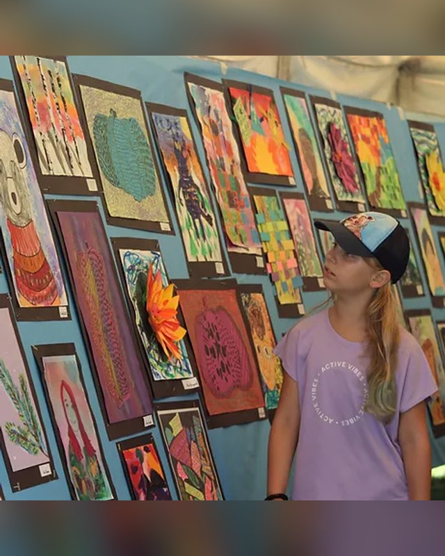 Artworks on display panels at an outdoor art show, with a young girl in a cap looking at them.