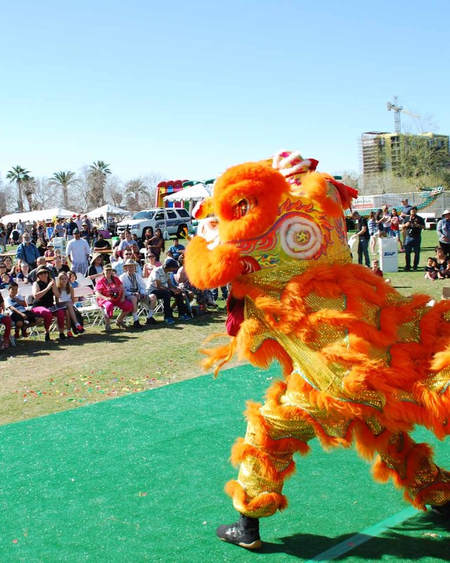 Crowd of festival visitors on a sunny lawn watching a lion dance performer in orange costume on a green mat.