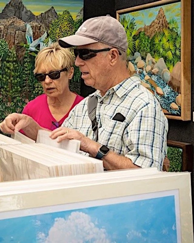 Two visitors sorting through stacked prints at an artist's booth, with mountain paintings on the wall behind them.