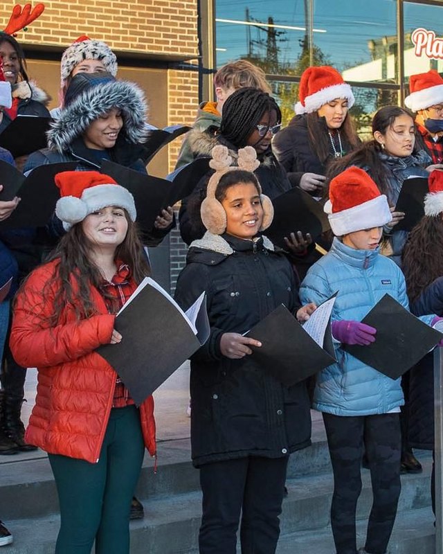 Children in Santa hats singing carols on steps outside a brick building at a holiday event.