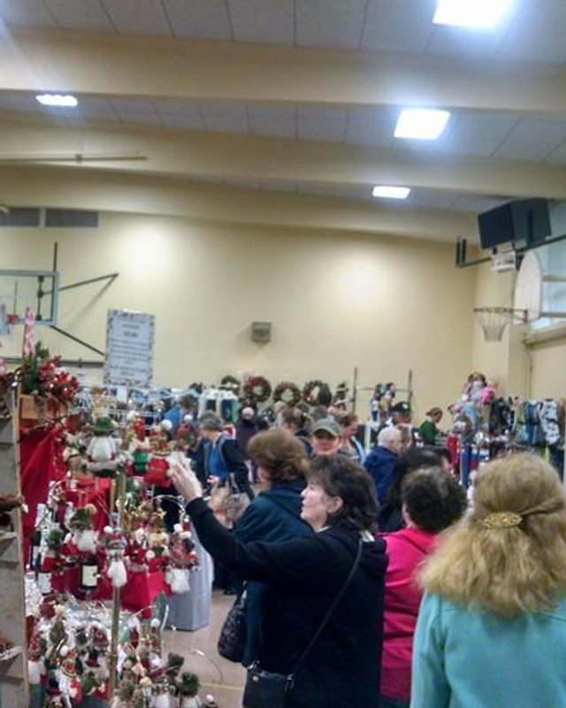 Crowd of shoppers browsing craft booths and holiday wreaths at an indoor fair in a school gym.