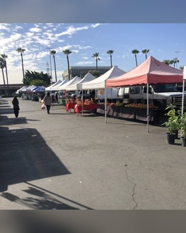 Outdoor market with white and red canopy tents, produce tables and shoppers on a paved lot under a blue sky.