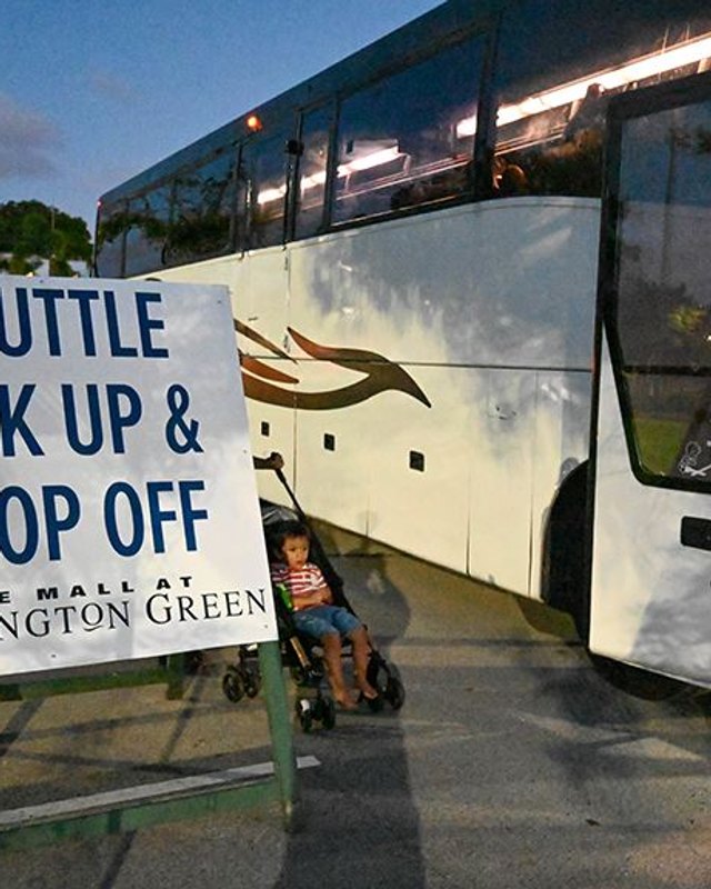 Shuttle bus beside a 'Shuttle Pick Up & Drop Off' sign at Wellington Green, with a small child in a stroller nearby.