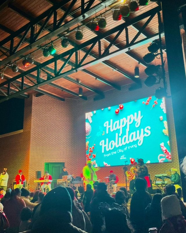 Crowd gathered at a holiday live music event with a Happy Holidays screen and string lights overhead.