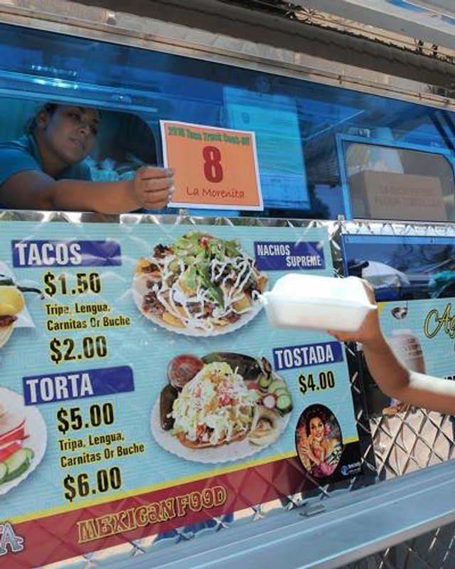 Taco truck service window with a number 8 sign, a menu board listing tacos, tortas and tostadas, and a hand holding a foam container from outside.