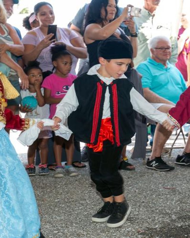 A boy in a black Greek costume with red trim dances among spectators at the Port Charlotte Greek Fest street festival.