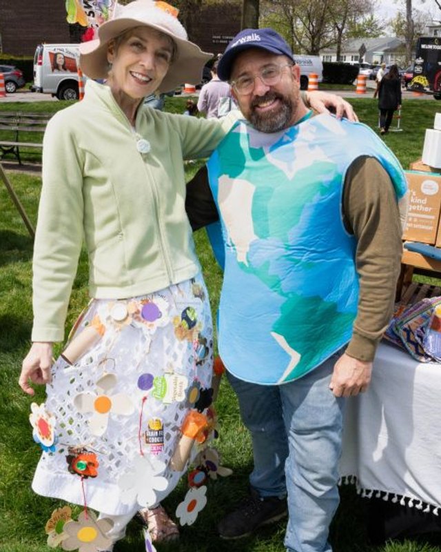 Two people in costume posing in front of a craft booth at the Norwalk Earth Day Festival outdoors.