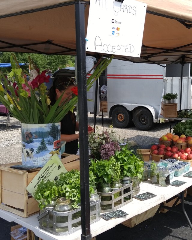 Vendor tent at the Manahawkin Flea Market with fresh flowers, peaches and tomatoes, and jars of preserves on a table, with a person partly visible behind the display.
