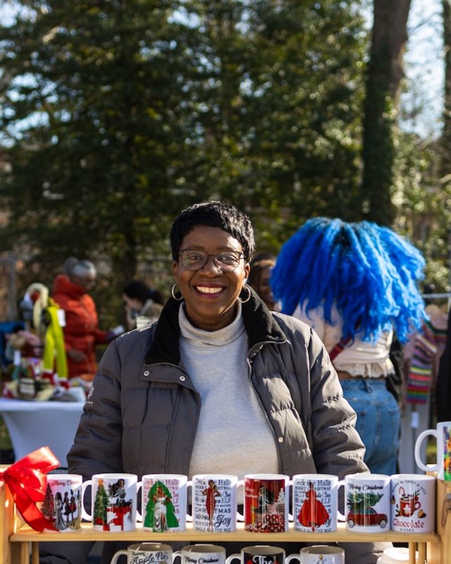 A smiling vendor behind a table of holiday-printed mugs at an outdoor market, with a blue-wigged figure and other people in the background.