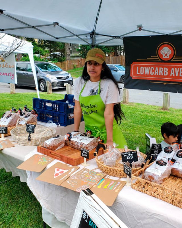 Vendor in a green 'Wheaton Farmers' apron behind a table of baked goods and packaged foods at a market booth under a white canopy, with a LowCarb Avenue banner.