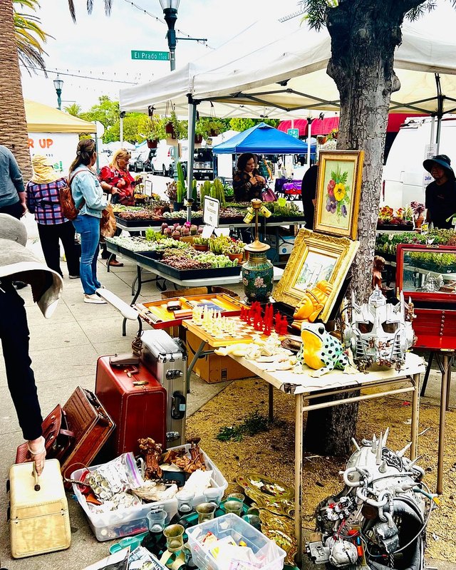 Market booths with antiques, framed art and vintage goods under white tents, with shoppers browsing.