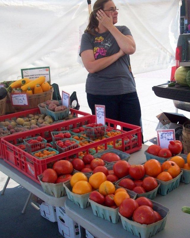 Vendor standing behind a produce stand with crates of berries, baskets of tomatoes and yellow squash at a farmers market.