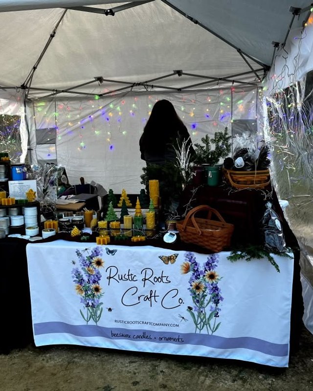 Craft vendor booth under a white tent with candles, jars, baskets and a Rustic Roots Craft Co cloth, with string lights overhead.