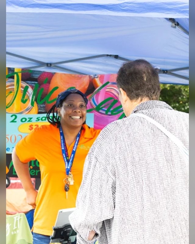 Market vendor in an orange shirt and lanyard at a colorful booth talking with a customer under a blue-and-white canopy.