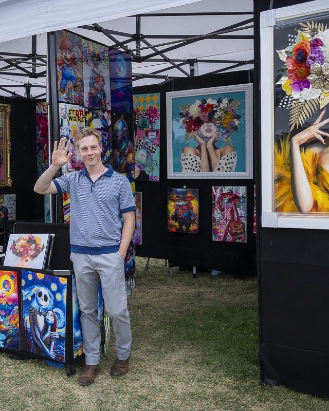 Artist standing in a Summerfest booth hung with colorful framed paintings under a white canopy.