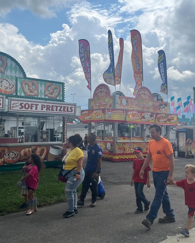 Families walk past a Soft Pretzels stand and other fair food booths with flags overhead at the Lorain County Fair.
