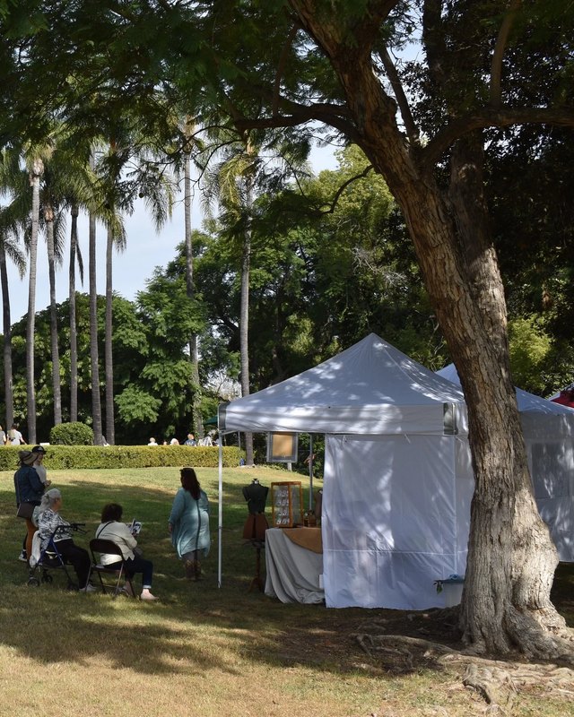 White vendor tents on a shaded lawn under palms and a large tree, with people seated near the tents.