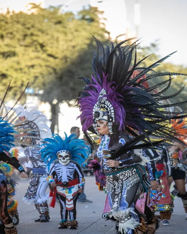 Costumed dancers in colorful feathered headdresses performing in a street parade for the Dallas Día de los Muertos festival