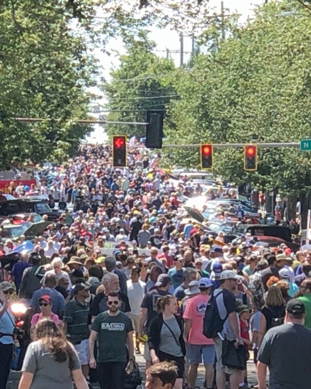 Large crowd walking a tree-lined street past tents and parked cars at the Greenwood Car Show