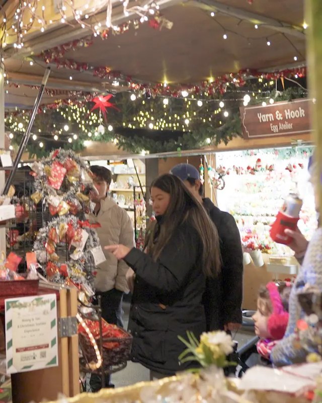 Shoppers browsing a decorated holiday market booth with string lights and wreaths.