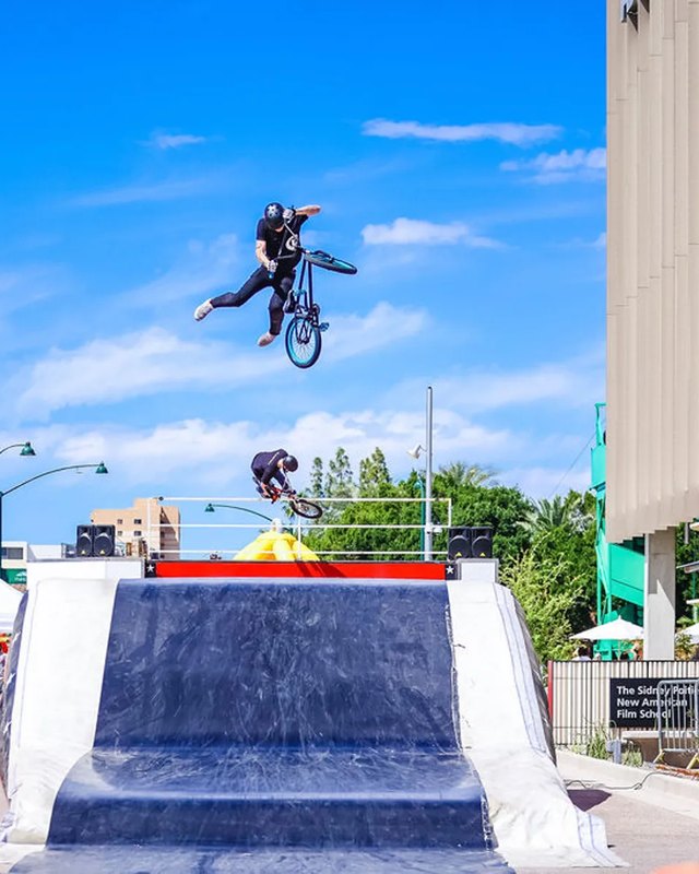 BMX rider mid-air over a stunt ramp on a plaza lined with palm trees in Downtown Mesa.