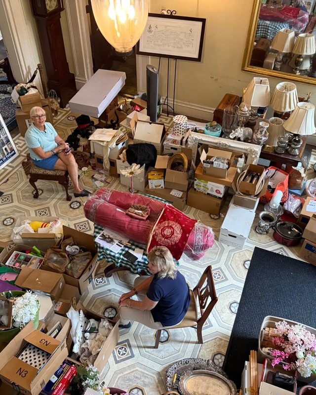 Two women sit among boxes, lamps and antiques piled on a patterned floor in a mansion room with a red rug.