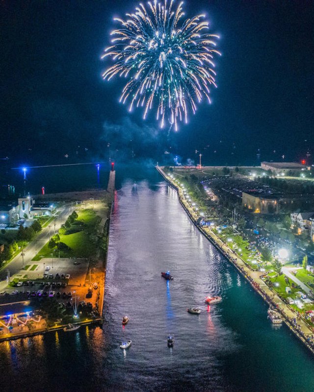 Fireworks burst over the harbor at night with a waterfront promenade lit up below, seen from above.