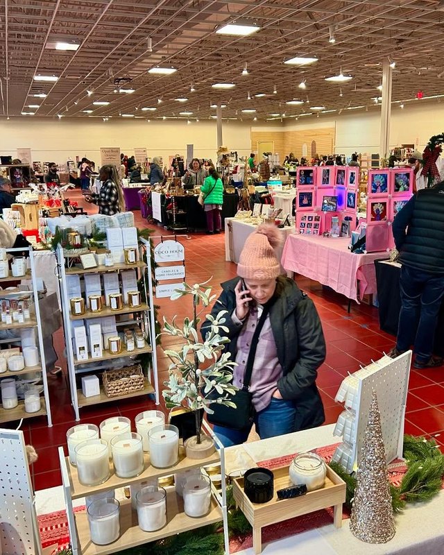 Indoor holiday market with candle and gift booths and shoppers browsing tables under ceiling lights.