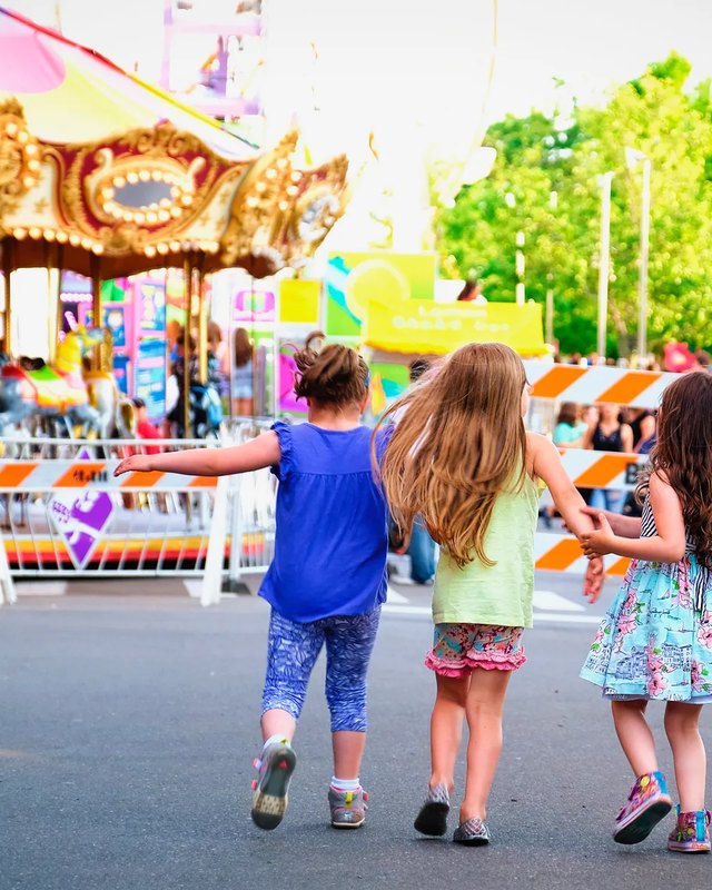 Three young girls walk away from the camera past a carousel and food signs at an outdoor fair.