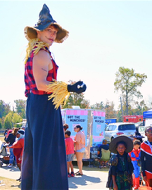 Costumed scarecrow character beside tents and families at an outdoor fall festival