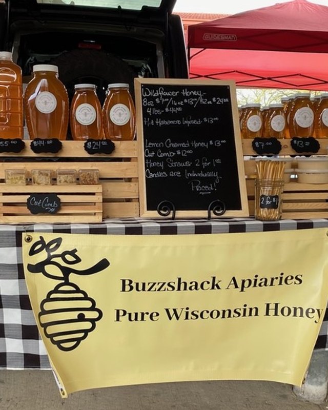 Buzzshack Apiaries honey booth with jars and crates of honey under a red canopy at the West Allis Farmers Market