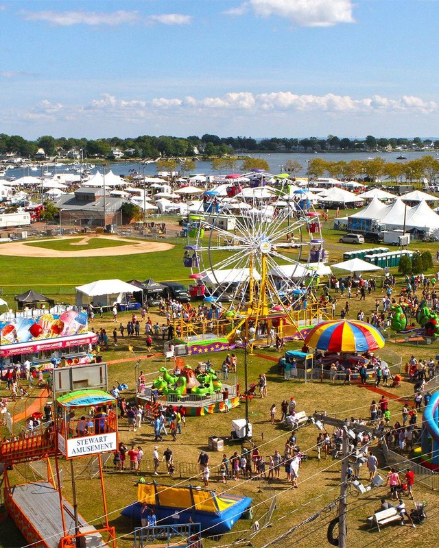 Aerial view of a festival crowd with tents, booths and amusement rides on a grassy park by the water