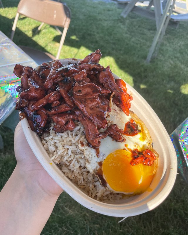A hand holding a rice bowl topped with braised meat and a fried egg, on a grassy lawn.