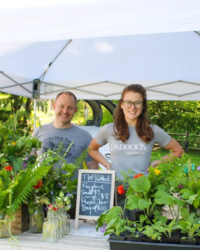 Two smiling vendors behind a plant stand with a chalkboard price sign at an outdoor market booth.